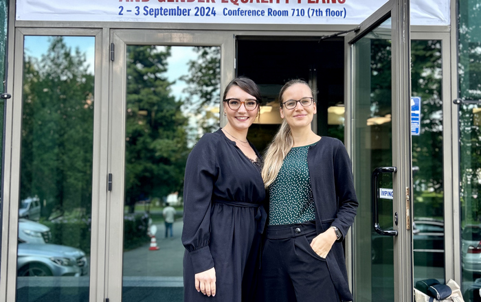 Zwei Frauen stehen vor einer geöffneten Glastür über der ein weißes Banner mit dem Konferenznamen und -datum in rot und blau hängt.